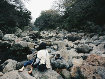 Rear view of woman sitting on rock at riverbank in forest