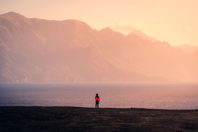 Remote back view of unrecognizable female explorer standing along rocky coast near sea on background of sunset sky and highlands on gran canaria