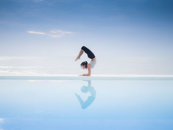 Full length of mature woman practicing yoga at poolside against sea and sky