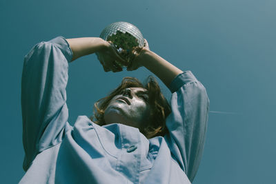 Low angle view of woman with glitter on face holding disco ball against clear sky