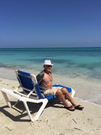 Mature man wearing hat relaxing on deck chair at beach against clear blue sky during sunny day