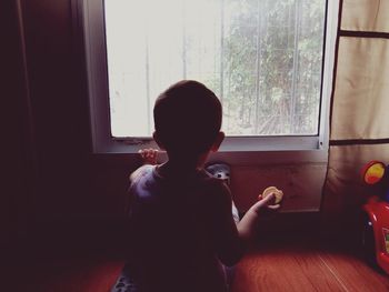 Rear view of boy looking through window while sitting at home