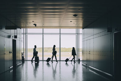 Side view of silhouette business colleagues pulling luggage while walking in corridor at airport