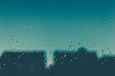 Silhouette buildings against blue sky seen through wet window in rainy season