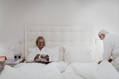 Happy senior woman having coffee while man standing by bed in hotel room