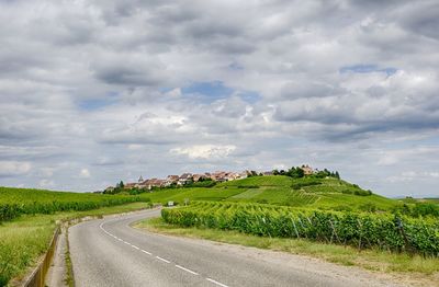 Road passing through agricultural field against cloudy sky