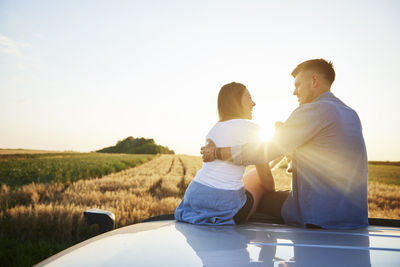 Couple sitting on car roof against sky