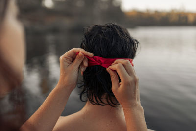 Crop anonymous female tourist tying red blindfold on head of partner against rippled water during trip