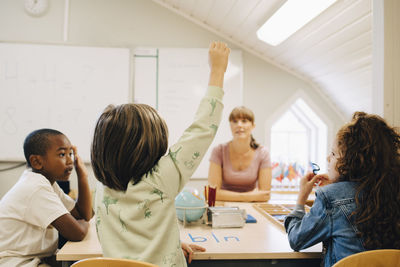 Schoolboy raising hand while answering teacher in classroom at school