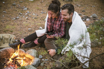 High angle view of happy couple wrapped in blanket while using mobile phone by fire pit at campsite