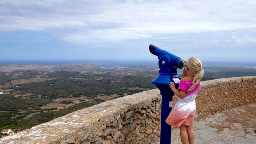 Girl looking through coin-operated binoculars against sky