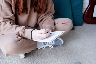 Young caucasian red-haired woman learning to play acoustic guitar at home, making notes 