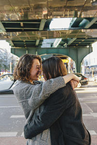 Cheerful lesbian couple embracing while standing on street under bridge in city