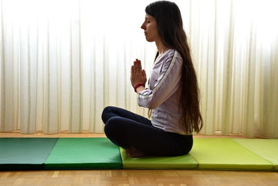 Woman meditating while sitting on exercise mat against curtains at home