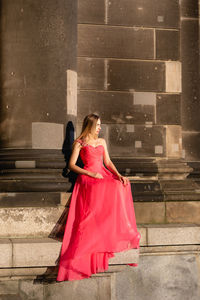 Full length of young woman in pink evening gown sitting against columns