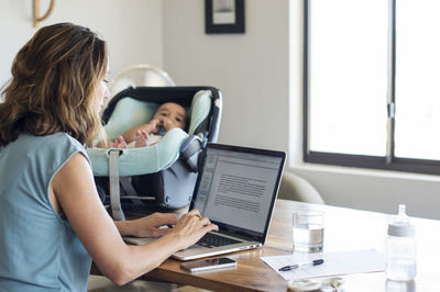 Mother using laptop computer by baby boy relaxing in seat on table at home