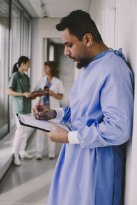 Side view of male surgeon writing on clipboard in hospital corridor