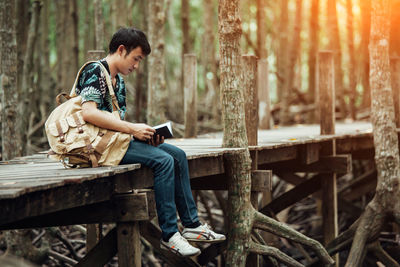 Side view of young man holding book while sitting on footbridge in forest