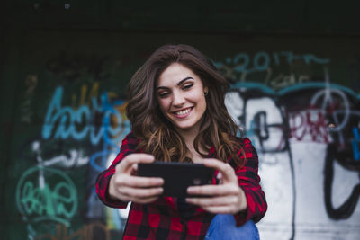 Smiling woman taking selfie through mobile phone against graffiti wall