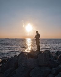 Man standing on rocks at sea shore against sky during sunset