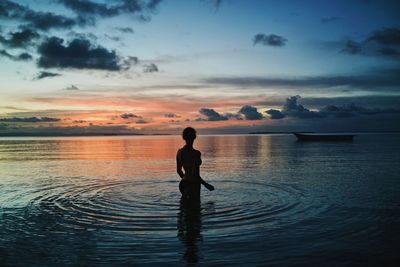 Silhouette boy standing in sea against sky during sunset