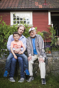 Portrait of man sitting with father and son against house in back yard