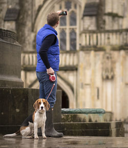 Man with dog photographing building through mobile phone on footpath