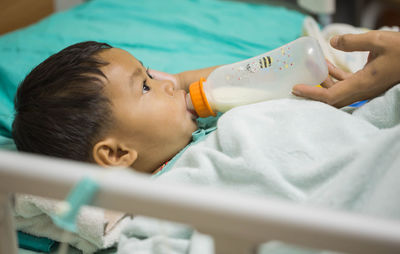 Cropped hands of parent feeding milk to son lying on bed in hospital