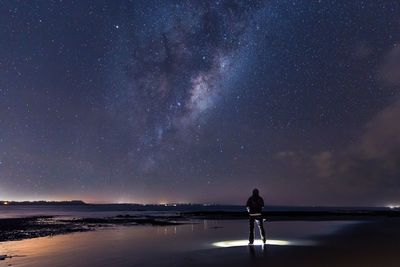 Rear view of man standing with illuminated flashlight on shore against milky way