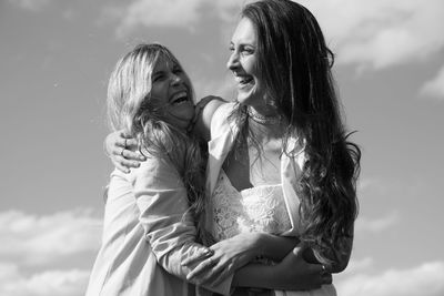 Cheerful young friends embracing while standing outdoors against sky