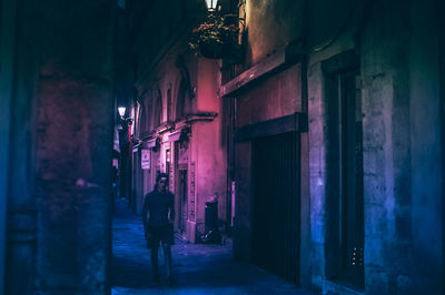 Rear view of man walking on illuminated street amidst buildings at night