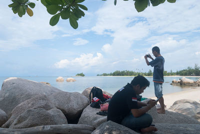 Friends sitting on rock by sea against sky