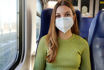 Happy young woman wearing medical mask sitting on train looking at camera.