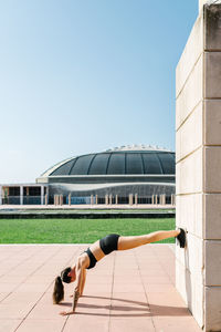 Full body side view of unrecognizable athletic female in sportswear doing feet on wall push ups near concrete building on city square