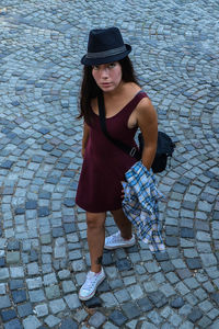 A young brunette woman in a burgundy dress and a black hat stands on the pavement road