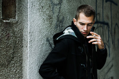 Young man smoking while standing against wall