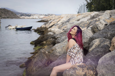 Side view of a young woman in summer dress sitting on the rocky coast of the sea