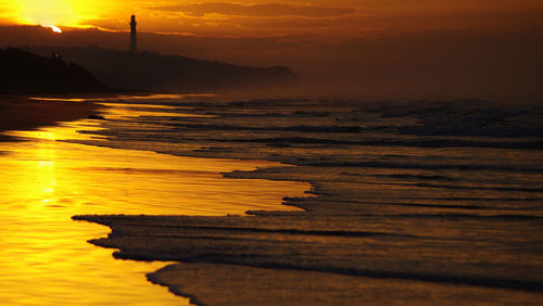 Deep orange sunset behind lighthouse reflected on beach with sea spray