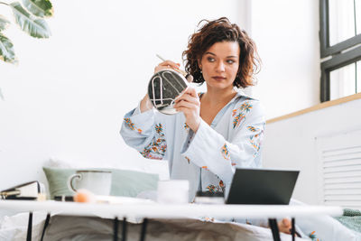 Young brunette woman in blue pajamas applies makeup in front of mirror sitting on bed at home