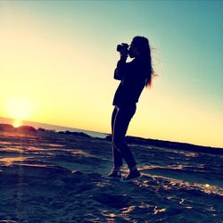 Side view of silhouette woman photographing at beach against clear sky during sunset
