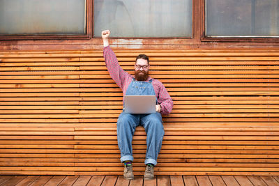 Cheerful brutal bearded hipster man working on laptop sitting on bench rising hand up celebrating.