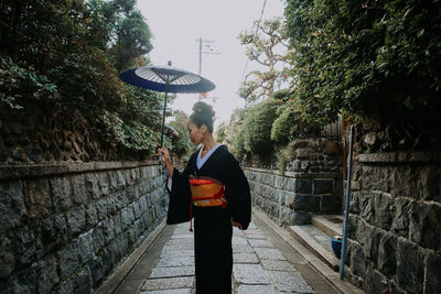 Mature woman holding umbrella standing by plants amidst alley