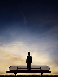 Low angle view of silhouette boy standing on bench against sky during sunset