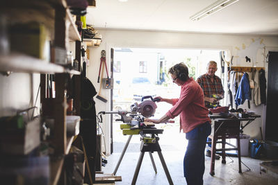 Side view of senior woman using circular saw while man holding drill in background