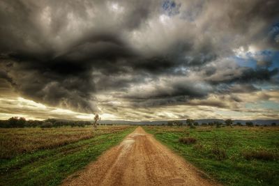 Dirt road passing through field against storm clouds