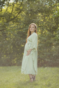 Young pregnant girl stands in park, looks at camera, green trees on background. brown haired woman