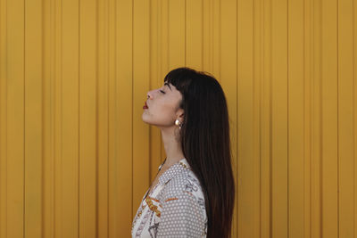 Side view of stylish young ethnic female with long dark hair in trendy dress standing against yellow wall on street and closed eyes thoughtfully