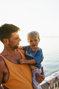 Father carrying cute son while standing on pier over lake against clear sky during sunset