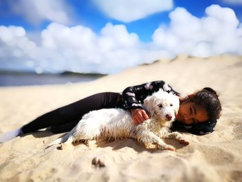 Teenage girl lying with dog on sand at beach against sky during sunny day
