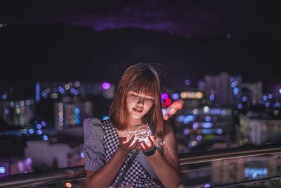 Woman holding illuminated string light against cityscape at night